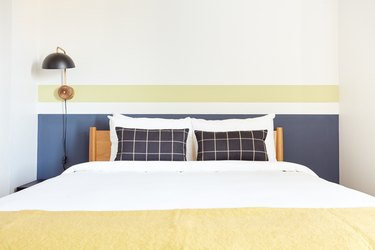 Modern bedroom with light wood headboard, checked pillows, yellow blanket, black metal sconce.